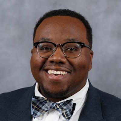 Smiling man in glasses wearing a navy suit, white shirt, and plaid bow tie. Background is a soft gray, conveying a formal and cheerful tone.
