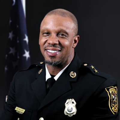 Uniformed police officer smiling, wearing a black formal jacket with insignia and badges. An American flag is visible in the dark background.
