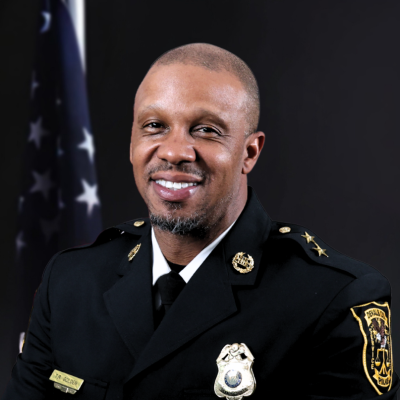 Uniformed police officer smiling, wearing a black formal jacket with insignia and badges. An American flag is visible in the dark background.