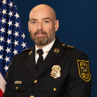 A man in a formal police uniform stands confidently against a dark blue background. An American flag is visible to the left. The tone is authoritative.