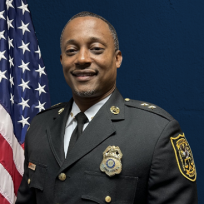 A man in a formal police uniform stands smiling in front of a U.S. flag and a dark blue background, conveying a sense of authority and pride.