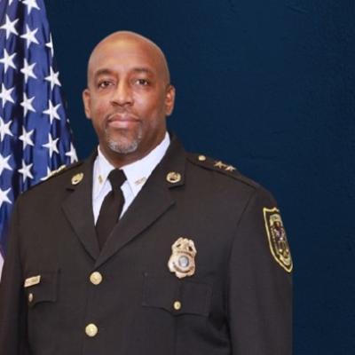 A person in a dark police uniform stands confidently beside an American flag. The blue background and badges suggest authority and professionalism.
