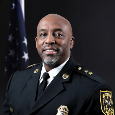 Man wearing a black traditional dress uniform with a gold badge, gold buttons, and a gold pin on either collar in front of the American flag and a light tan background.