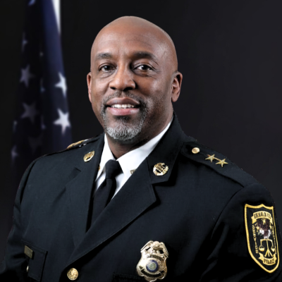 Man wearing a black traditional dress uniform with a gold badge, gold buttons, and a gold pin on either collar in front of the American flag and a light tan background.