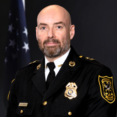 Image of man wearing a black traditional police officer uniform with a gold badge, gold buttons, and a gold pin on either collar in front of the American flag in the background