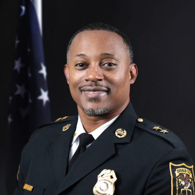 Man wearing a black traditional dress uniform with a gold badge, gold buttons, and a gold pin on either collar in front of the American flag and a black background.