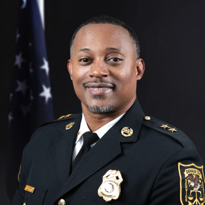 Man wearing a black traditional dress uniform with a gold badge, gold buttons, and a gold pin on either collar in front of the American flag and a black background.