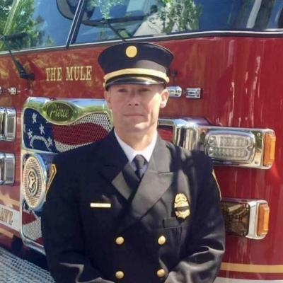 A firefighter in formal uniform stands in front of a bright red fire truck named "The Mule." The scene conveys pride and professionalism.
