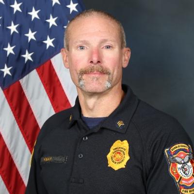 A firefighter in uniform stands solemnly in front of an American flag. The badge on his shirt features a fire department emblem, conveying duty and honor.