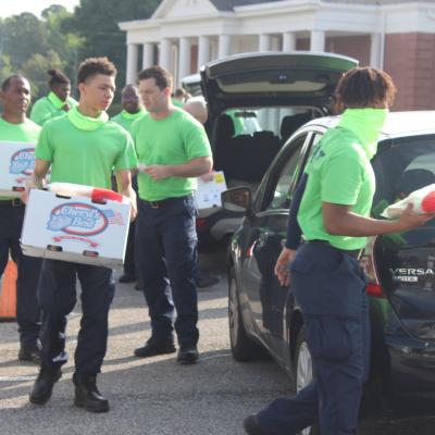 Volunteers in bright green shirts unload boxes from a car during a community event. The scene is busy, with a collaborative and energetic mood.