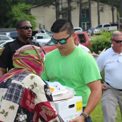 A man in a green shirt and sunglasses hands a box of supplies to a person in a colorful headscarf at an outdoor community event, surrounded by volunteers.