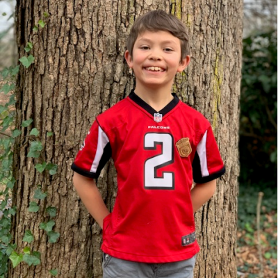 A smiling child in a red football jersey with the number 2 stands against a large tree trunk in a wooded area, conveying a cheerful and playful mood.