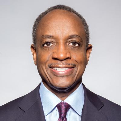 A man in a dark suit with a light blue shirt and patterned tie smiles warmly against a plain light gray background. The image conveys a professional tone.