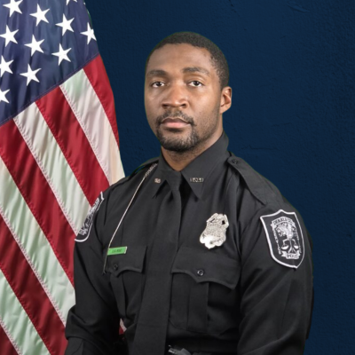 Uniformed police officer posing against a dark blue background, standing beside an American flag. The tone is serious and professional.