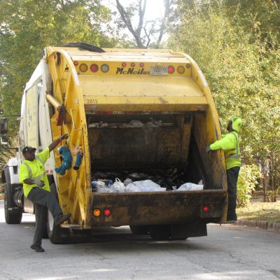 Two sanitation workers in high-visibility attire collect trash from a yellow garbage truck on a tree-lined residential street.