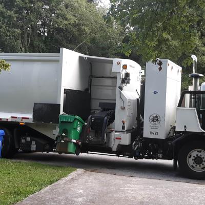 A garbage truck lifts a green bin to empty its contents. A worker in a yellow vest operates the truck. A blue recycling bin is visible nearby, surrounded by leafy trees.