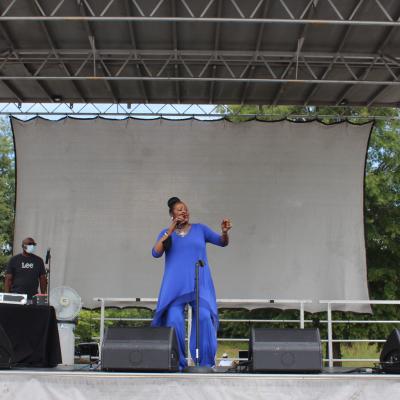 A woman in a vibrant blue outfit energetically sings on an outdoor stage. Two men stand to the left near sound equipment. Trees are visible in the background.