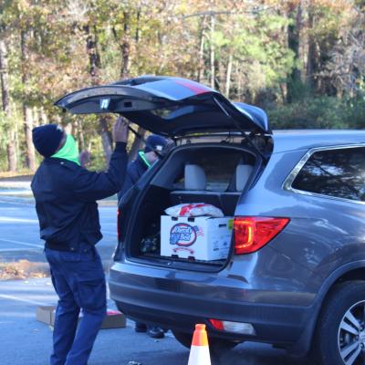 Person in a green scarf loads items into an open SUV trunk with a moving box. Surrounding cones and autumn trees create a busy, outdoor scene.