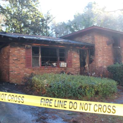 A brick house with fire damage has charred walls, broken windows, and a burnt-out car. Yellow "Fire Line Do Not Cross" tape surrounds the scene.
