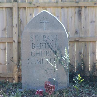 A stone monument with a cross and the words "St. Paul Baptist Church Cemetery" stands in front of a wooden fence, surrounded by overgrown plants.