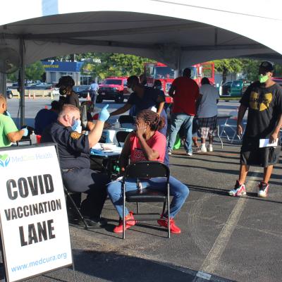 Outdoor COVID vaccination site under a tent, where a healthcare worker in uniform administers a vaccine to a seated woman. Sign in foreground.