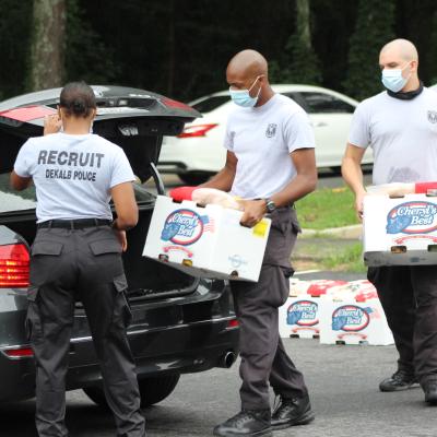 Four police recruits wearing masks load boxes labeled "Cheryl's Best" into a car trunk. The scene is cooperative, with a sense of community service.