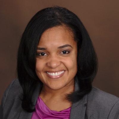 A woman with shoulder-length black hair smiles warmly. She is wearing a gray blazer over a purple shirt against a simple brown background.
