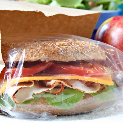 A whole grain sandwich with lettuce, sliced turkey, cheddar, and tomato in plastic wrap peeks from a brown bag, next to a shiny red apple on a table.