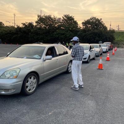 A line of cars waits in a parking lot, organized with orange traffic cones. A man in casual attire speaks to a driver. The sky is overcast.