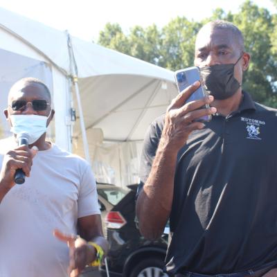 Two men wearing masks and sunglasses stand outdoors. One speaks into a microphone, while the other holds up a smartphone. A large tent is in the background.