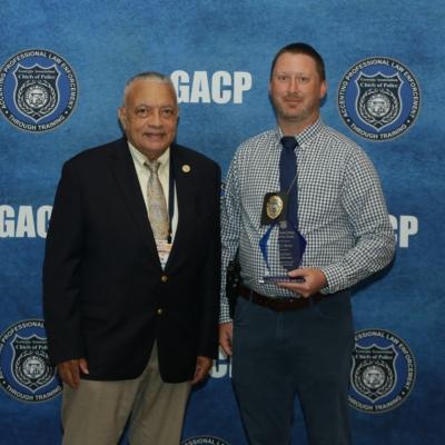 Two men stand smiling in front of a blue GACP backdrop. One man wears a suit, the other holds an award, wearing a checked shirt and tie.