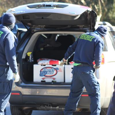 Two people in blue uniforms with "RECRUIT" on the back load supplies into an SUV. A large box labeled "Cheryl's Best" is seen. Scene is outdoors.