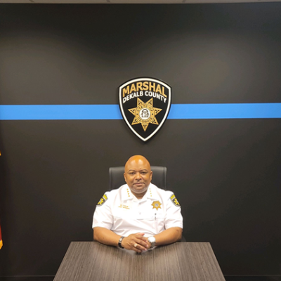 Person in a white uniform sits at a desk, flanked by US and Georgia flags, under a DeKalb County Marshal badge. The room feels official and formal.