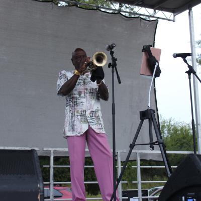 A musician in colorful attire plays the trumpet on an outdoor stage, with a mic and a tablet on stands nearby, conveying a vibrant, live performance.