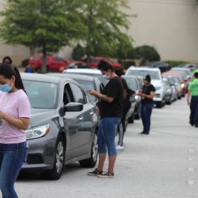 People wearing masks stand beside a line of parked cars, engaging with drivers. The scene depicts a drive-thru event on an overcast day.