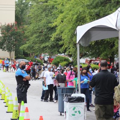 People wearing masks line up outside under trees next to a building, supervised by a police officer. Cones guide them, hinting at an organized event.