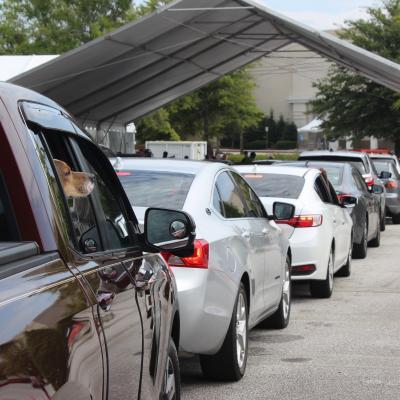 A line of cars waits under a large tent at an outdoor event. A dog looks out from a truck window, adding a curious and eager vibe to the scene.