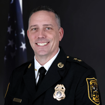 Man wearing a black traditional dress uniform with a gold badge, gold buttons, and a gold pin on either collar in front of the American flag and a black background.