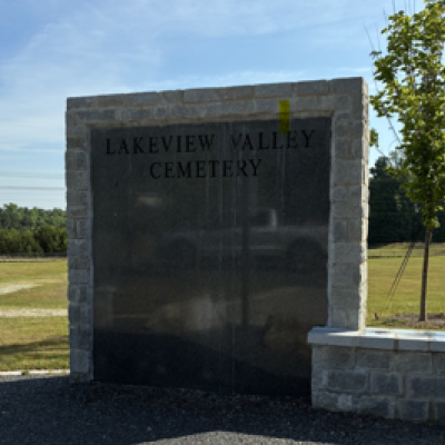 Image of the Lakeview Cemetery headstone with a rock in front of it and trees and the cemetery land behind it.