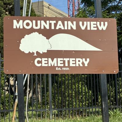 Image of a brown and white sign with Mountain View Cemetery written on it with a tree and mountain sketch in the middle. Sign is mounted on a black gate in the grass with building behind it.
