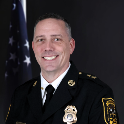 Headshot of a smiling male police official in uniform, standing in front of a dark background with an American flag to the left.