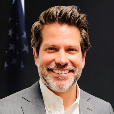 Image of a smiling man standing in front of a dark background with an American flag wearing a grey suit jacket and a white button up shirt. 