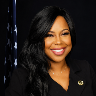 Professional portrait of a woman with long dark wavy hair, wearing a black wrap dress with a DeKalb County pin, in front of an American flag