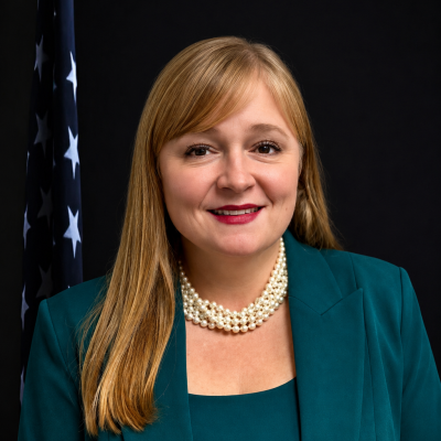 Professional portrait of a woman with long blonde hair, wearing a teal blazer and pearl necklace, in front of an American flag