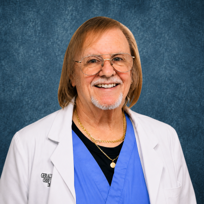 Professional portrait of a man with shoulder-length blonde hair, glasses, and a gray goatee, wearing a white lab coat over blue scrubs, in front of a blue textured background.