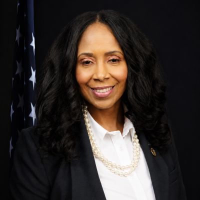 Professional portrait of a woman with curly dark hair, wearing a pearl necklace, white collared shirt, and black blazer with a DeKalb County pin, in front of an American flag.