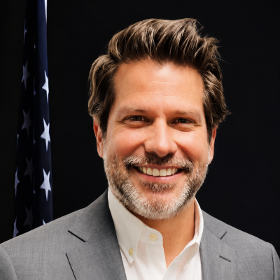 Professional portrait of a man with brown and gray hair and beard, wearing a gray blazer and light collared shirt, in front of an American flag
