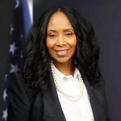 Professional headshot of a woman with curly dark hair, wearing a black blazer, white collared shirt, and pearl necklace, smiling in front of an American flag.
