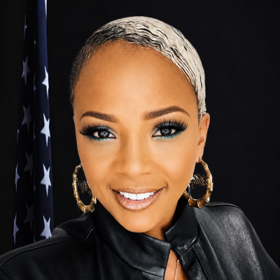 Professional portrait of a woman with short platinum-blonde and black hair, wearing gold hoop earrings and a black leather jacket, in front of an American flag