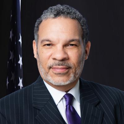 Official portrait of a man with gray curly hair and a gray beard, wearing a dark pinstriped suit with a purple tie and white shirt, standing in front of an American flag against a dark background.